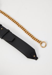 Zwarte leren band met gouden klinknagels naast een dikke gouden ketting met een ronde sluiting op een witte achtergrond.