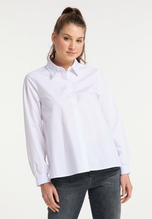 Giovane donna con capelli castani raccolti, che indossa una camicia bianca con bottoni e jeans scuri su uno sfondo chiaro e uniforme.