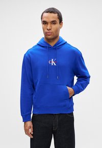 Férfi élénk kék Calvin Klein kapucnis pulóverben, elülső zsebbel és fekete farmerral, egy sima fehér háttér előtt áll.