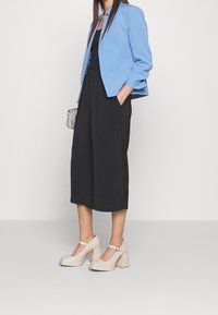 Modrý blazer s krátkými rukávy, černé culottes a béžové boty na blokovém podpatku s páskem kolem kotníku. Texturovaná kabelka dodává detail.