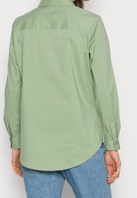 En kvinna ses bakifrån i en ljusgrön långärmad skjorta tillsammans med blå jeans, vilket framhäver skjortans krage och manschetter.