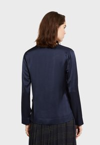 Mornarsko plava satenska bluzica s dugim rukavima, glatke teksture, zaobljenim rubom i jednostavnim dizajnom. Nosila se preko crne karirane suknje.