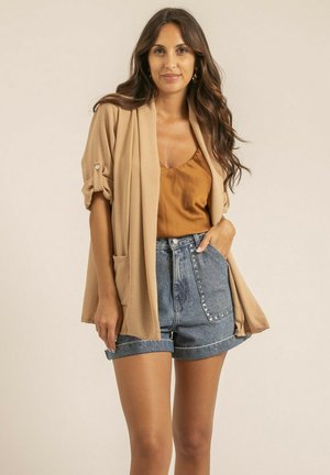 Femme aux longs cheveux bruns ondulés portant un cardigan ouvert couleur sable, un débardeur couleur rouille et un short en jean taille haute avec des revers et des détails à clous.