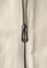 Nærbillede af en metal lynlås skyder på lyst beige stof, delvist lynet med synlige sammenkædede tænder og syningsdetaljer.