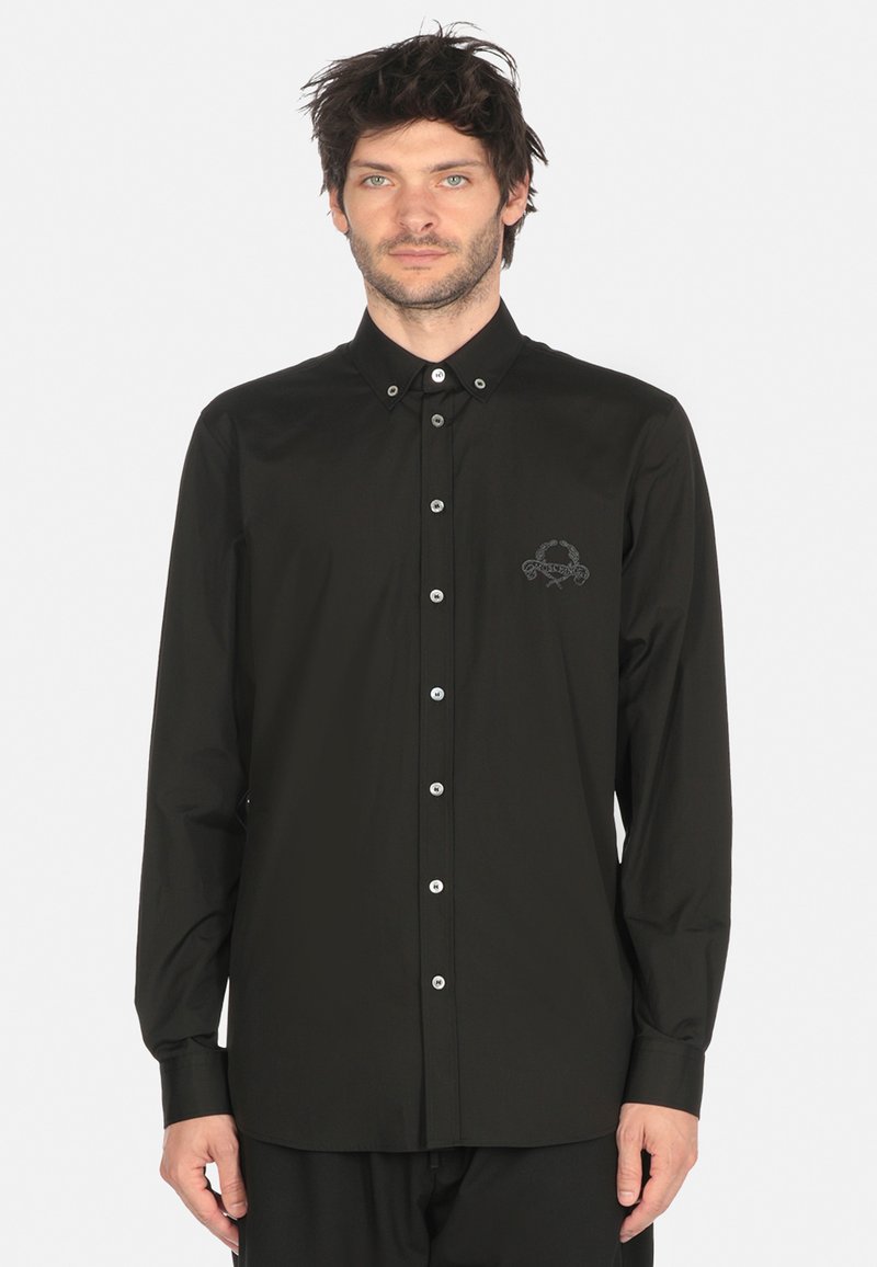 Uomo che indossa una camicia nera con bottoni e maniche lunghe, con un piccolo stemma ricamato sul lato sinistro del petto, in piedi davanti a uno sfondo semplice.