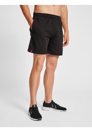 Homme portant un short de sport noir et des chaussures de course noires, debout devant un fond clair uni, une main dans la poche.