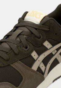 Gros plan d'une basket ASICS marron et beige avec des lacets texturés, des panneaux en suède et en mesh, et une étiquette de languette de marque.
