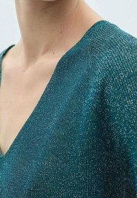 Miętowy sweter z dekoltem w kształcie litery V, wykonany z połyskującej, teksturowanej dzianiny. Zbliżenie pokazuje dekolt oraz subtelny efekt połysku.