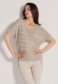 Beige gebreide trui met korte mouwen, open geweven textuur, brede halslijn en een ontspannen pasvorm, gecombineerd met lichtbeige strakke broek.