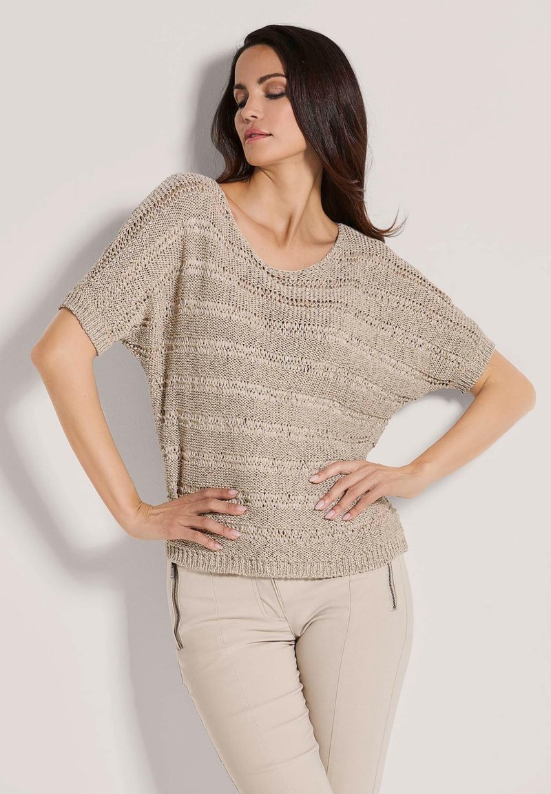 Beige gebreide trui met korte mouwen, open geweven textuur, brede halslijn en een ontspannen pasvorm, gecombineerd met lichtbeige strakke broek.