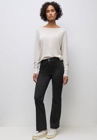 Donna con capelli ricci in piedi indossa una maglia beige a maniche lunghe, pantaloni neri aderenti e scarpe da ginnastica bianche su uno sfondo grigio chiaro.