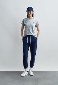 Kobieta w szarej koszulce, granatowych spodniach dresowych z białym sznurkiem, białych tenisówkach i granatowej czapce, stojąca z rękami w kieszeniach.