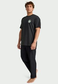 Mann, der barfuß steht und ein schwarzes T-Shirt mit einem kleinen runden Grafik sowie schwarze hochgekrempelte Hosen trägt, vor einem schlichten Hintergrund.
