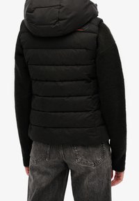 Svart pufferväst med horisontellt quiltad design, fastsatt huva och kontrasterande stickade ärmar. Matcher med bleka grå jeans.