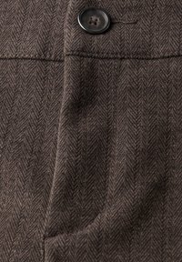 Brunt herringbone-mönstrad tyg med en svart knapp, featuring en sidficka och texturerad yta. Skräddarsydd design med subtila detaljer.