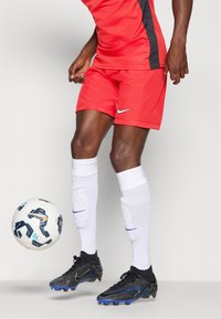 Rød Nike sportsdrakt og shorts, svarte detaljer, kombinert med hvite høye sokker og svarte fotballsko, med en mønstret fotball i nærheten.