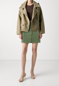 Jupe en velours côtelé vert avec boutons sur le devant, associée à une veste en simili cuir vert olive et un haut court marron, complétée par des talons transparents.