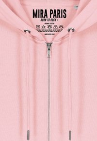 Sweat à capuche rose zippé avec cordons, fermeture éclair en métal, étiquette indiquant "MIRA PARIS BORN TO ROCK" et instructions de lavage à l'intérieur du col.