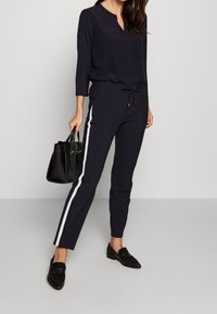 Navy blouse met een V-hals en twee borstzakken, gecombineerd met navy joggingbroeken met witte zijstrepen en een verstelbare tailleband. Een zwarte handtas maakt de look compleet.