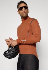 Maillot de cyclisme à manches longues marron en tissu Polartec, avec une fermeture éclair quart de zip et une poche noire. Un short de cyclisme noir et un casque sont tenus à la main.