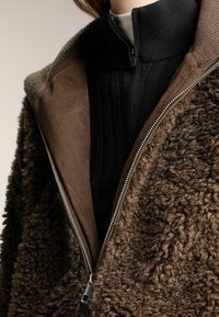 Bruine jas met rits, met een getextureerde, pluizige buitenkant en een gladde binnenvoering, met daaronder een geribd zwart shirt met hoge hals.