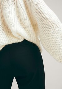 Osoba ubrana w gruby, kremowy sweter w stylu knit, włożony w wysokie, dopasowane czarne spodnie, widoczna od pasa w dół.