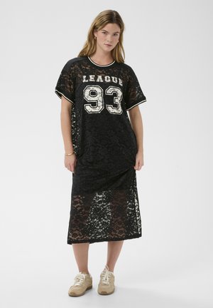 Žena oblečená v čiernych čipkovaných midi šatách s nápisom "LEAGUE 93" na prednej strane, doplnených béžovými teniskami, stojaca pred jednoduchým bielym pozadím.