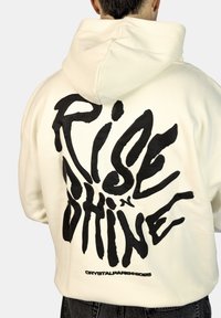 Felpa crema con design grafico nero, con il testo "Rise and Shine" sul retro. Tessuto morbido, vestibilità rilassata e cappuccio con cordino.