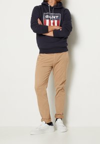 Sudadera con capucha azul marino con logotipo, pantalones chinos color beige y zapatillas blancas con detalles en azul marino. La sudadera tiene cordones; los chinos tienen puños enrollados.