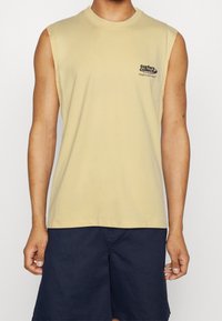 Man wearing a sleeveless beige shirt with "surfers collective" text and navy blue shorts, standing against a plain white background.