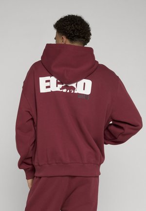 Osoba nosiaca bordovú mikinu s kapucňou a zodpovedajúce nohavice, ukazujúca chrbát s bielym nápisom "ECKO" a grafikou nosorožca na mikine.