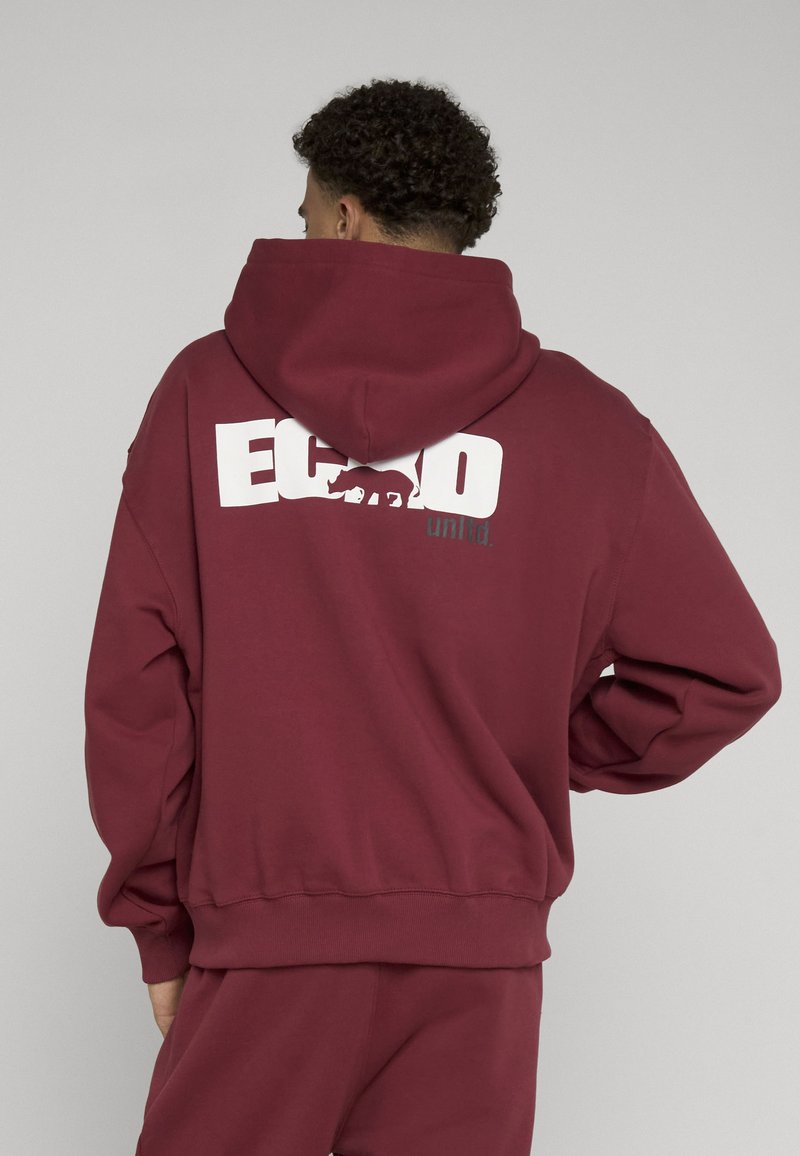 Personne portant un sweat à capuche bordeaux et un pantalon assorti, montrant le dos avec le texte blanc "ECKO" et un dessin de rhinocéros sur le sweat.