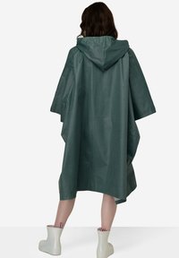 Grøn vandtæt poncho med hætte, der har et løst og bredt design. Lavet af glat, let materiale. Bæres med lyse støvler.