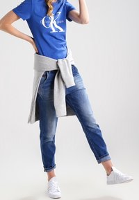 En kvinna i blå Calvin Klein t-shirt, uppvikta blå jeans, vita sneakers och en grå sweatshirt knuten runt midjan, poserar med ett ben lyft.