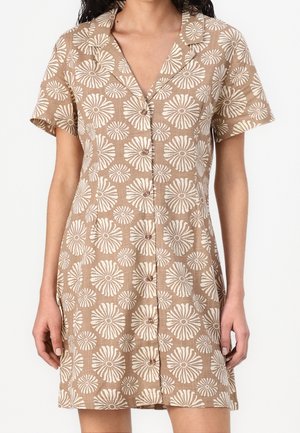 Kvinde iført en kortærmet brun skjortekjole med hvidt blomstermønster og forreste knapper, stående med armene afslappet ned langs siderne.