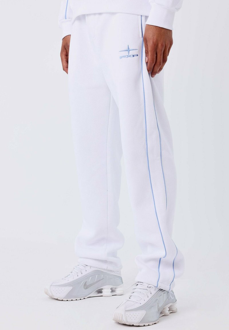 Close-up van de onderkant van het lichaam in witte joggingbroek met lichtblauwe zijstrepen en logo, gecombineerd met witte en grijze Nike sneakers.