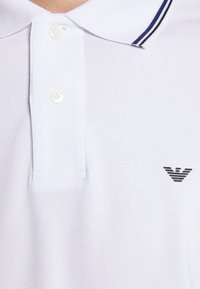 Polo blanc en tissu texturé, avec une patte de boutonnage à deux boutons et un col rayé noir et bleu. Logo noir sur la poitrine.