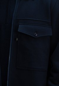 Navyblauwe zip-up jas met een textuurstof, een borstzak met een knoop en een klein logo-etiket aan de zijkant. Over het geheel een gladde afwerking.
