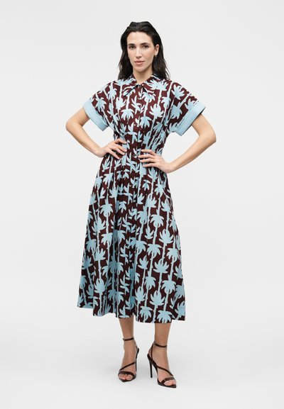 Γυναίκα με σκούρα μαλλιά φοράει καφέ midi φόρεμα με ανοιχτά γαλάζια τροπικά φυλλώματα και μαύρα ψηλοτάκουνα σανδάλια, στέκεται με τα χέρια στη μέση.