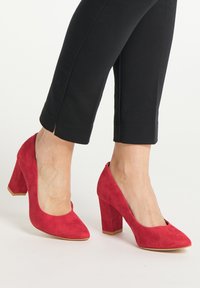 Picioare purtând pantofi cu toc înalt din nubuc roșu, asortați cu pantaloni scurți negri, pe un fond alb.