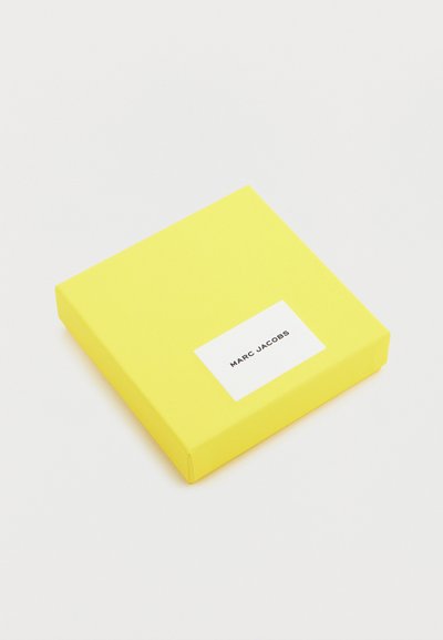 Κίτρινο τετράγωνο κουτί με λευκή ετικέτα που εμφανίζει το κείμενο "MARC JACOBS" σε μαύρο, τοποθετημένο σε απλό λευκό φόντο.