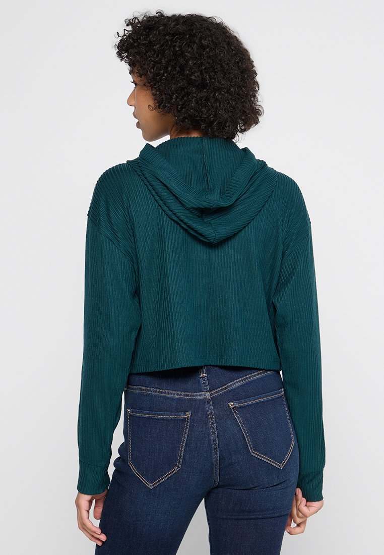 Sudadera corta de tejido de canalé en verde oscuro con capucha ajustable, que cuenta con mangas largas y se combina con jeans de mezclilla oscura.
