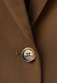 Nærbillede af en brun stofjakke med revers og en blank, firhullet knap, der er fastgjort gennem en syet knaphul.