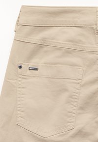 béžové bavlněné kalhoty s kapsou na zadní straně, se šikmým prošíváním a malou kovovou značkou poblíž kapsy. Hladká textura.