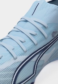Svetlo modni športni čevelj z teksturiranim pletenim zgornjim delom, ravnimi vezalkami in temno modrim ukrivljenim pasom ob strani.