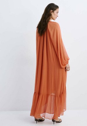 Femme aux longs cheveux foncés portant une robe maxi fluide orange brûlé et des talons noirs à lanières, debout devant un fond clair uni.