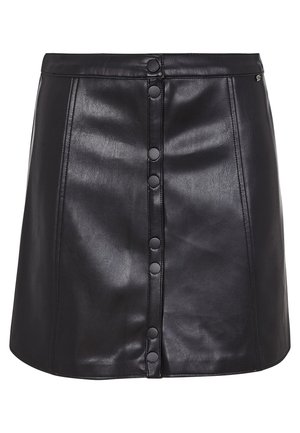 Femme portant un haut blanc oversize et une jupe en cuir noire boutonnée, tenant une veste en cuir noire assortie, portant des baskets et des chaussettes noires.