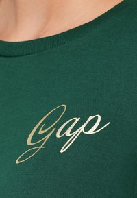 Camiseta de algodón verde con cuello redondo que presenta un logo en dorado con la palabra "Gap" en el pecho. Textura suave y diseño ajustado.