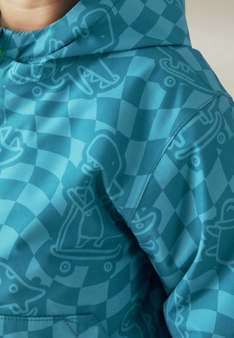 Veste à carreaux bleue avec des motifs de dinosaures dessinés en bleu clair chevauchant des skateboards visibles sur l'épaule et la poitrine.