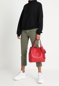 Červená kožená kabelka se strukturovaným tvarem a postranní kapsou. Model má na sobě černou mikinu a olivově zelené zkrácené kalhoty, které doplňuje bílými teniskami.
